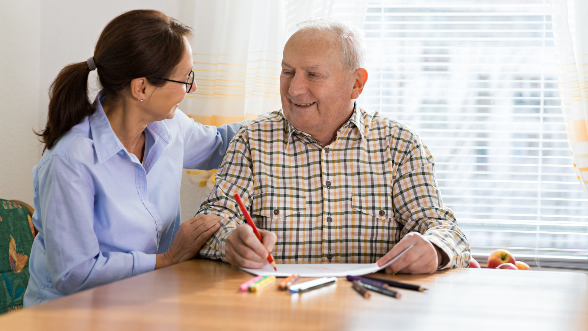 Woman with glasses smiles, puts arm on man's shoulder as he draws with pencil at a table.
