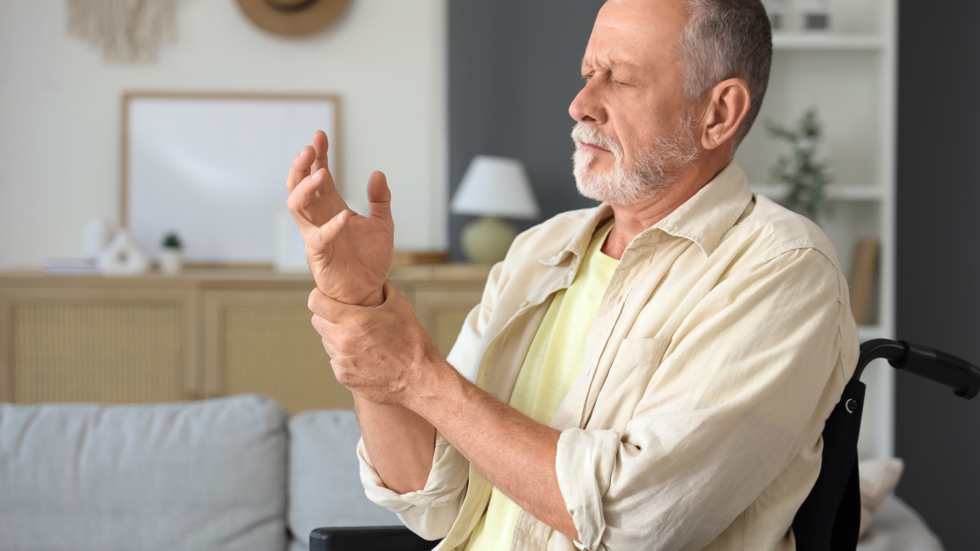 Older man in a wheelchair holding his wrist, looking down with a pained expression indoors.