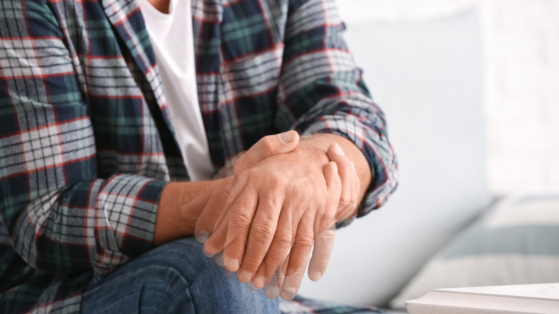 Man holding his wrist, possibly due to pain; wearing plaid shirt and jeans, seated indoors.