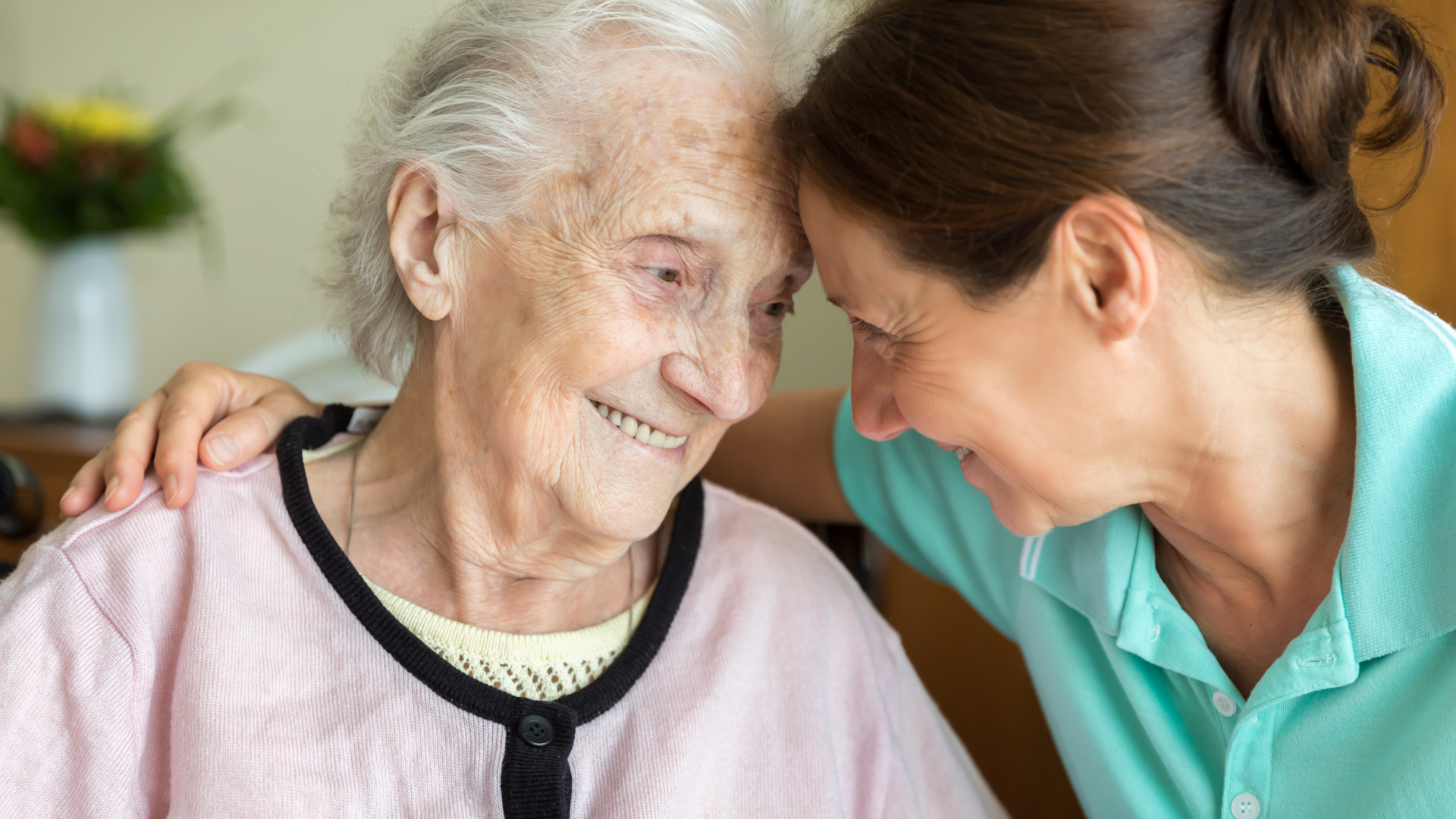 Woman comforts elderly woman, both smiling and touching foreheads.
