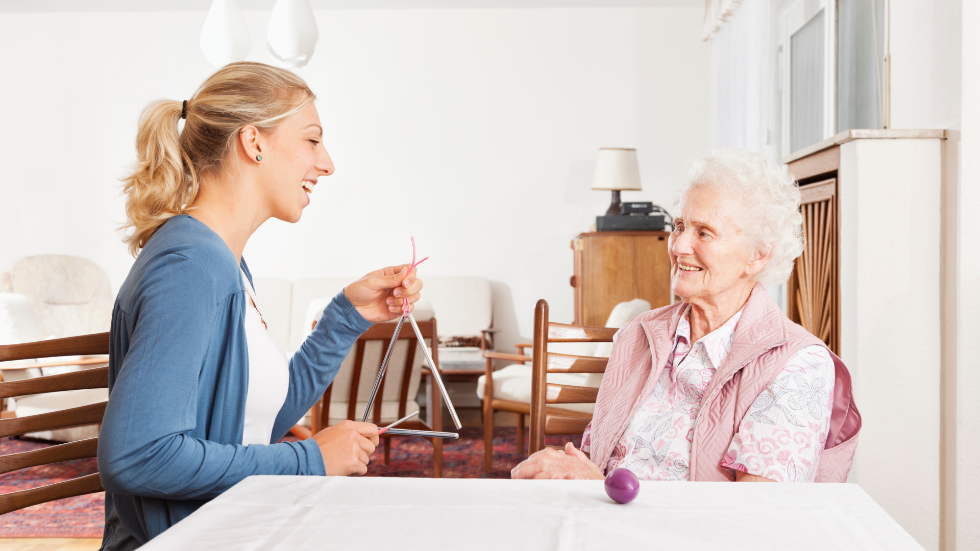 Woman holding a triangle, smiling at an elderly woman in a home.