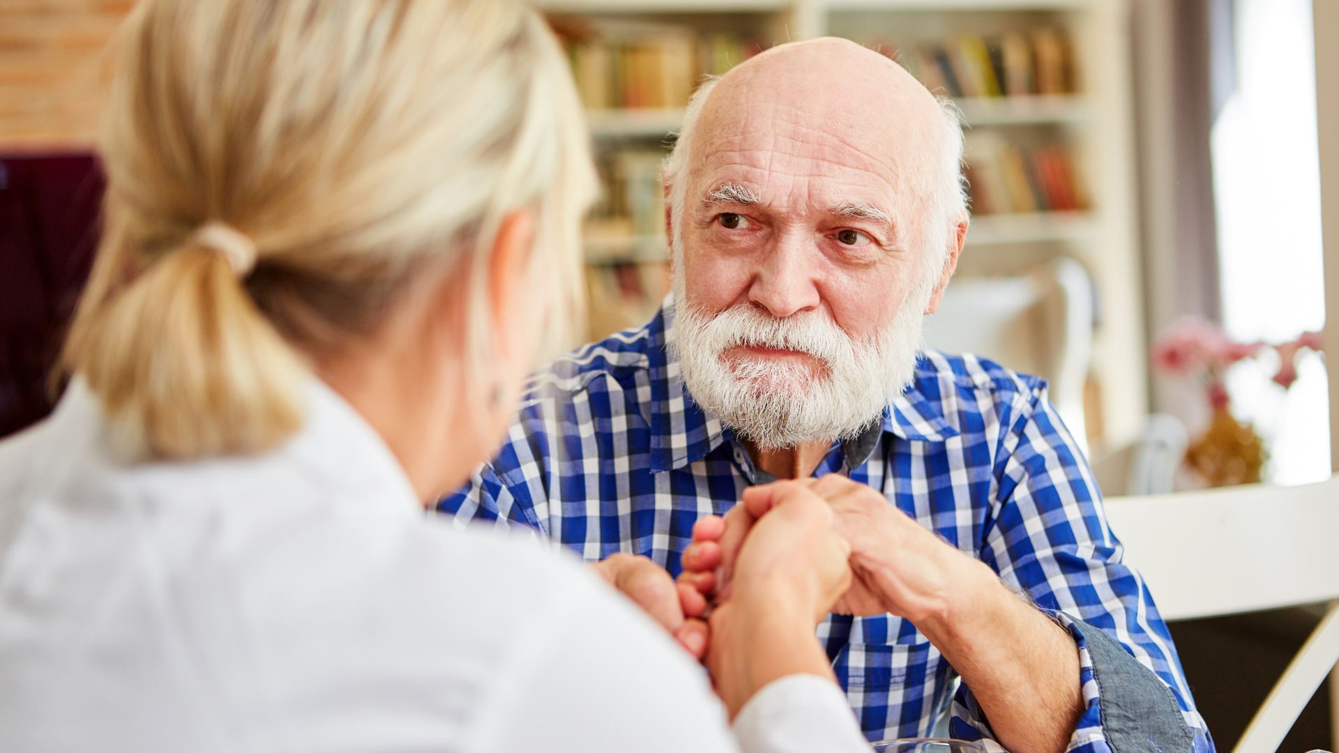 A senior man, with a white beard and blue plaid shirt, holds hands with a blonde woman.