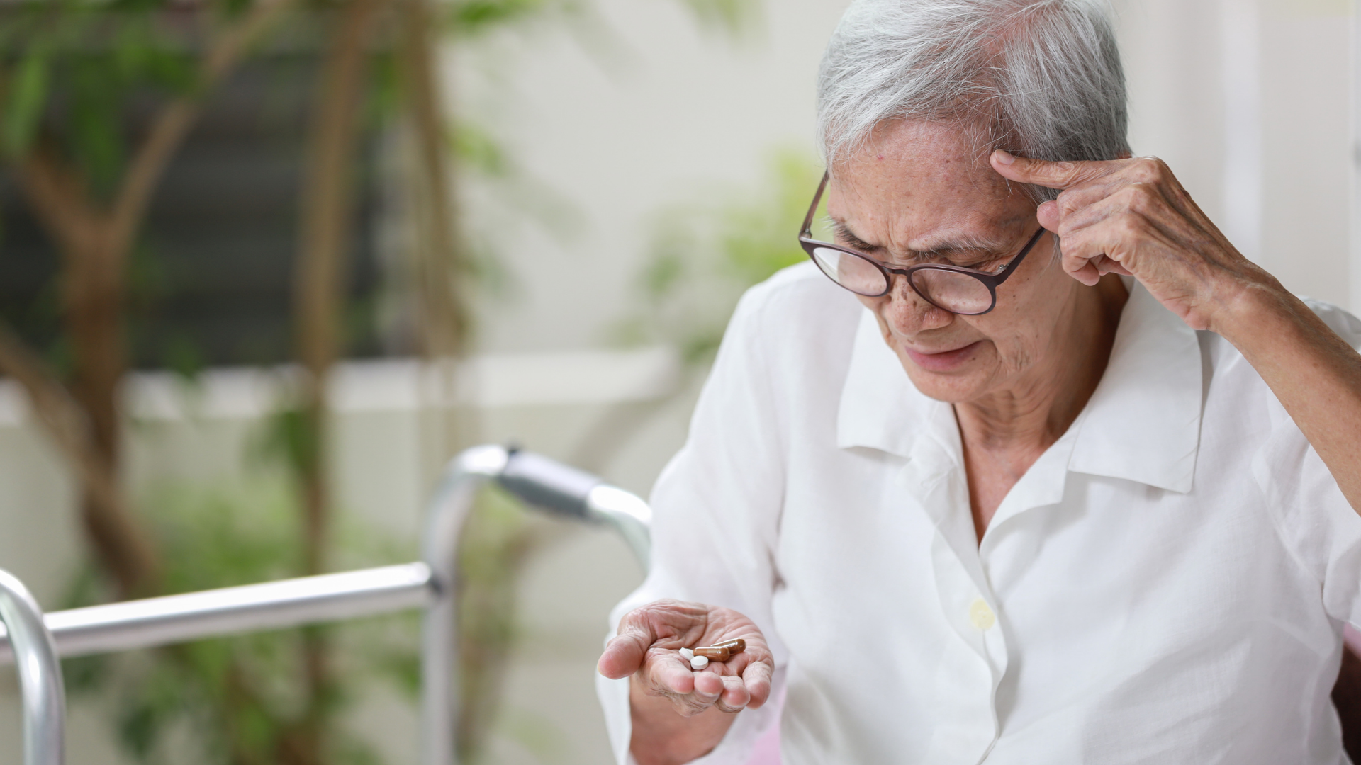 Elderly woman in glasses, holding pills, touching her head in thought; blurred background.