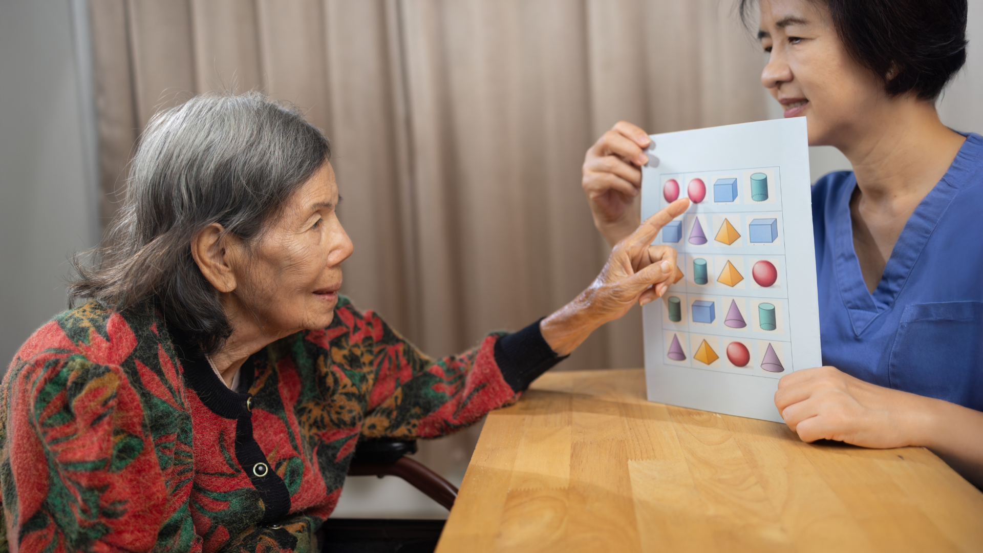 Elderly woman pointing at a chart with shapes and colors during a cognitive test with a healthcare worker indoors.