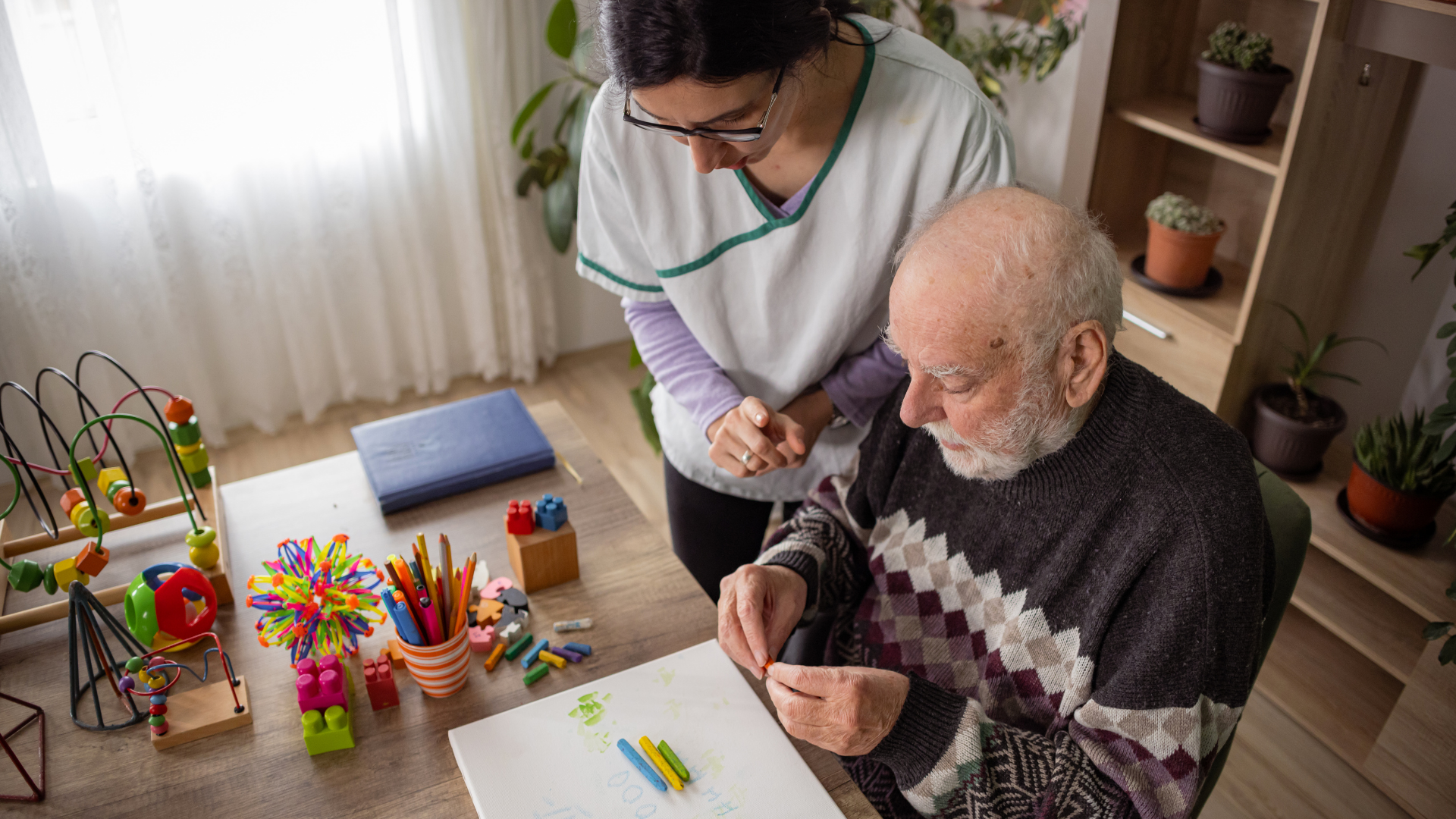 Caregiver assists an elderly man with a craft activity at a table, indoor setting.