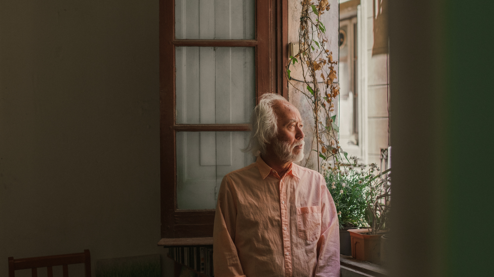 Elderly man with white hair and beard looks out a sunny window.
