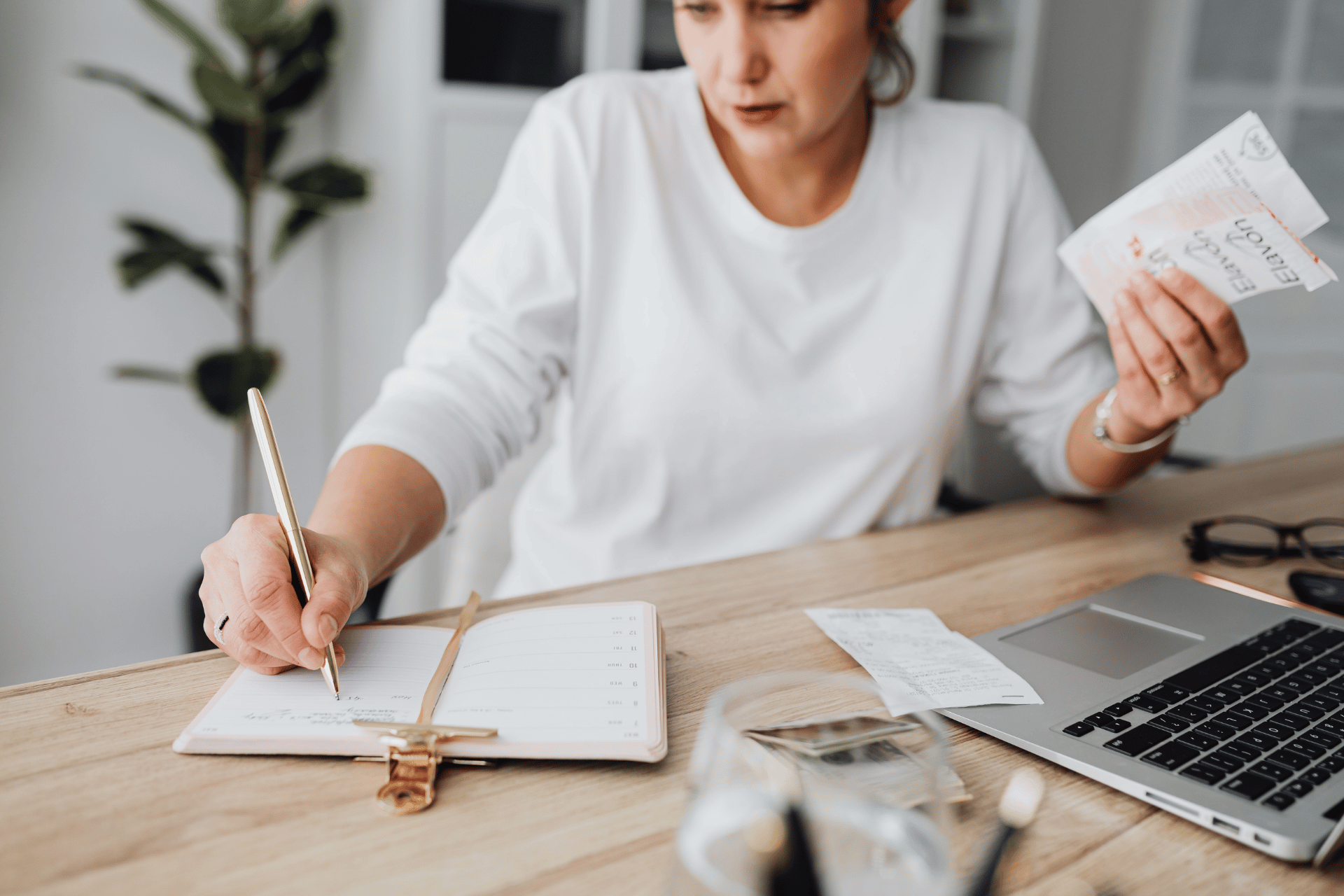 Woman writing in a planner while holding receipts at a desk with a laptop.