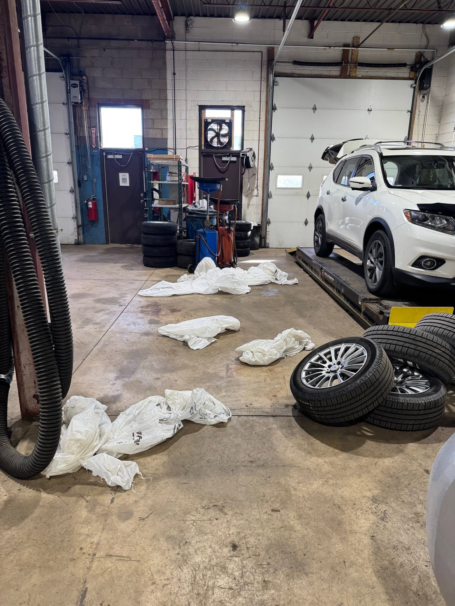 Auto repair shop with a white SUV on a lift, loose tires, and debris scattered on the concrete floor | SAI Automotive Inc.