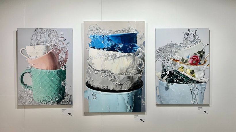 Drie abstracte schilderijen van koffiekopjes in pastel- en blauwtinten hangen aan een galeriemuur.