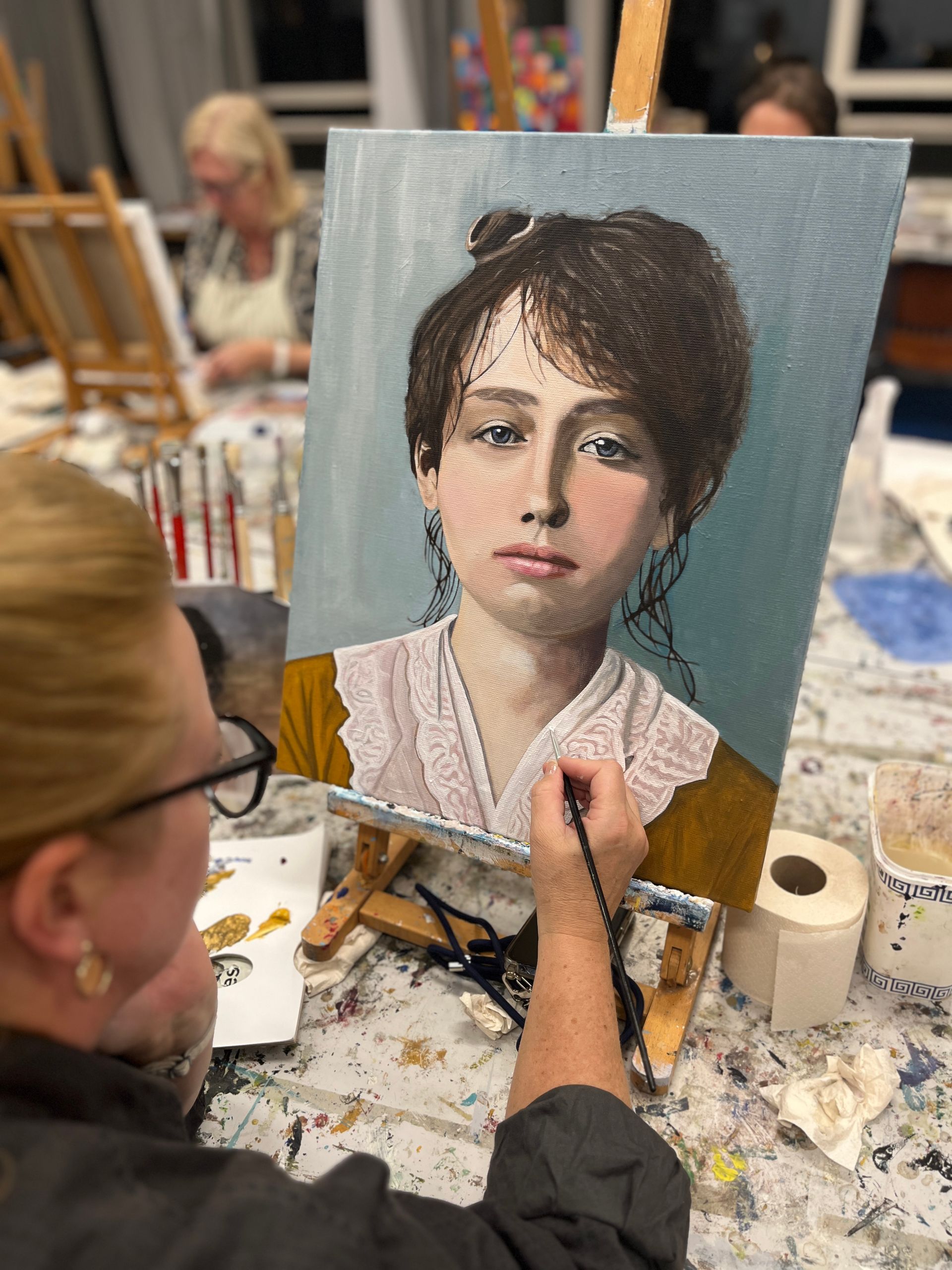 Een kunstenaar schildert een portret op een doek in een atelier, met een onderwerp in focus en de benodigdheden voor de verf zichtbaar.