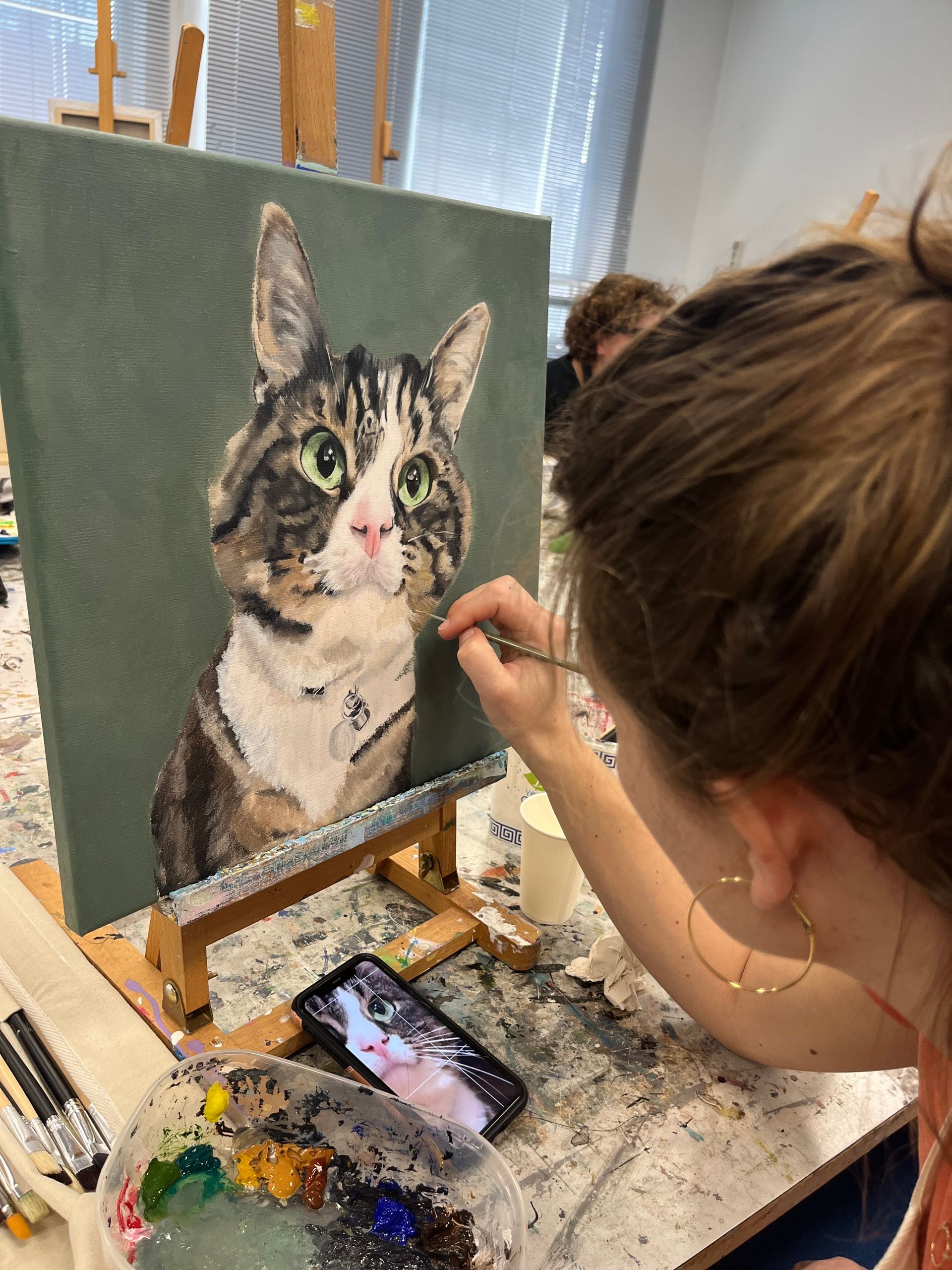 Een persoon schildert een kattenportret op canvas aan een schildersezel, met schildersbenodigdheden op tafel.