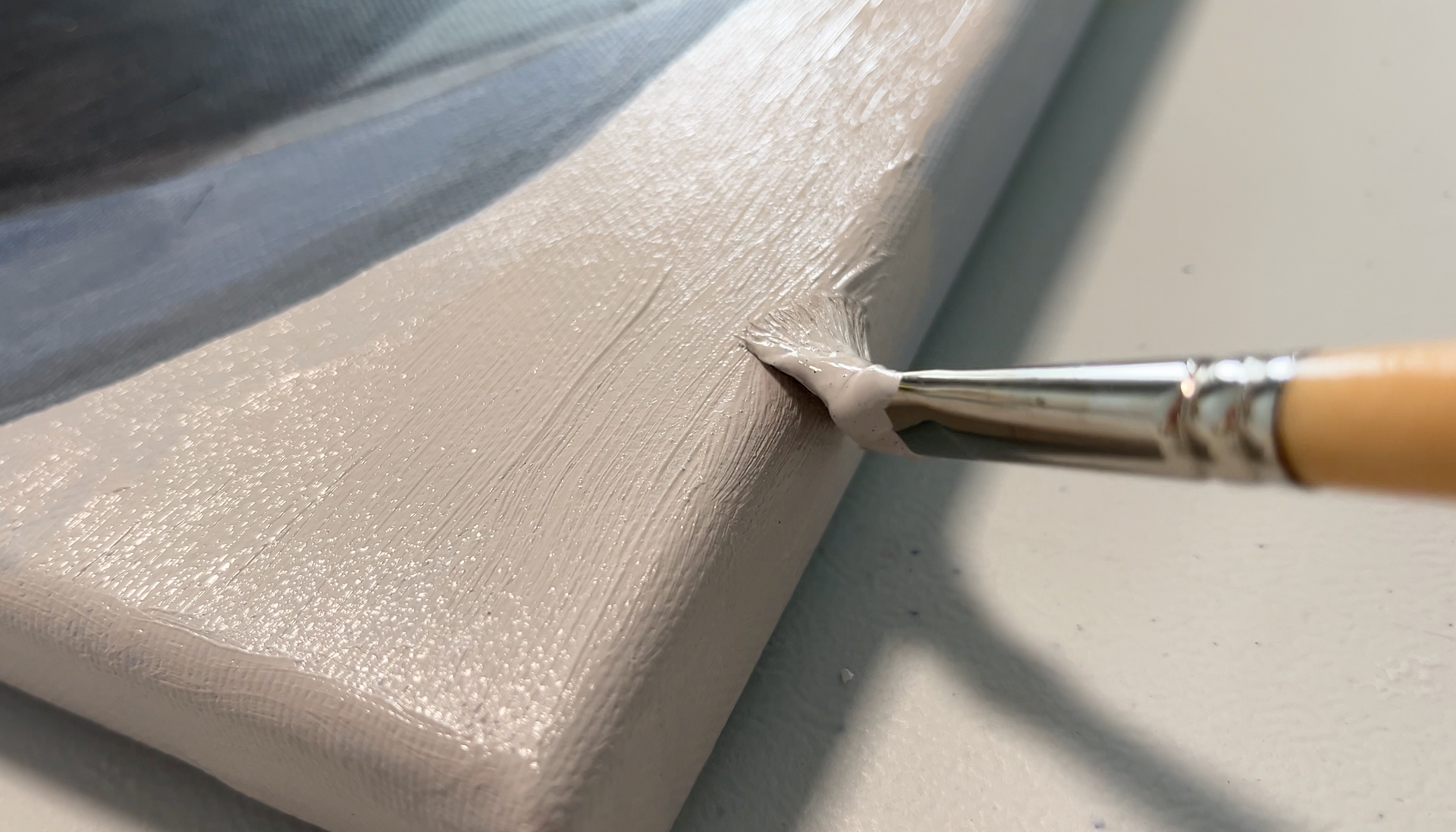 Een penseel raakt de hoek van een grijs, gestructureerd canvas op een tafel.