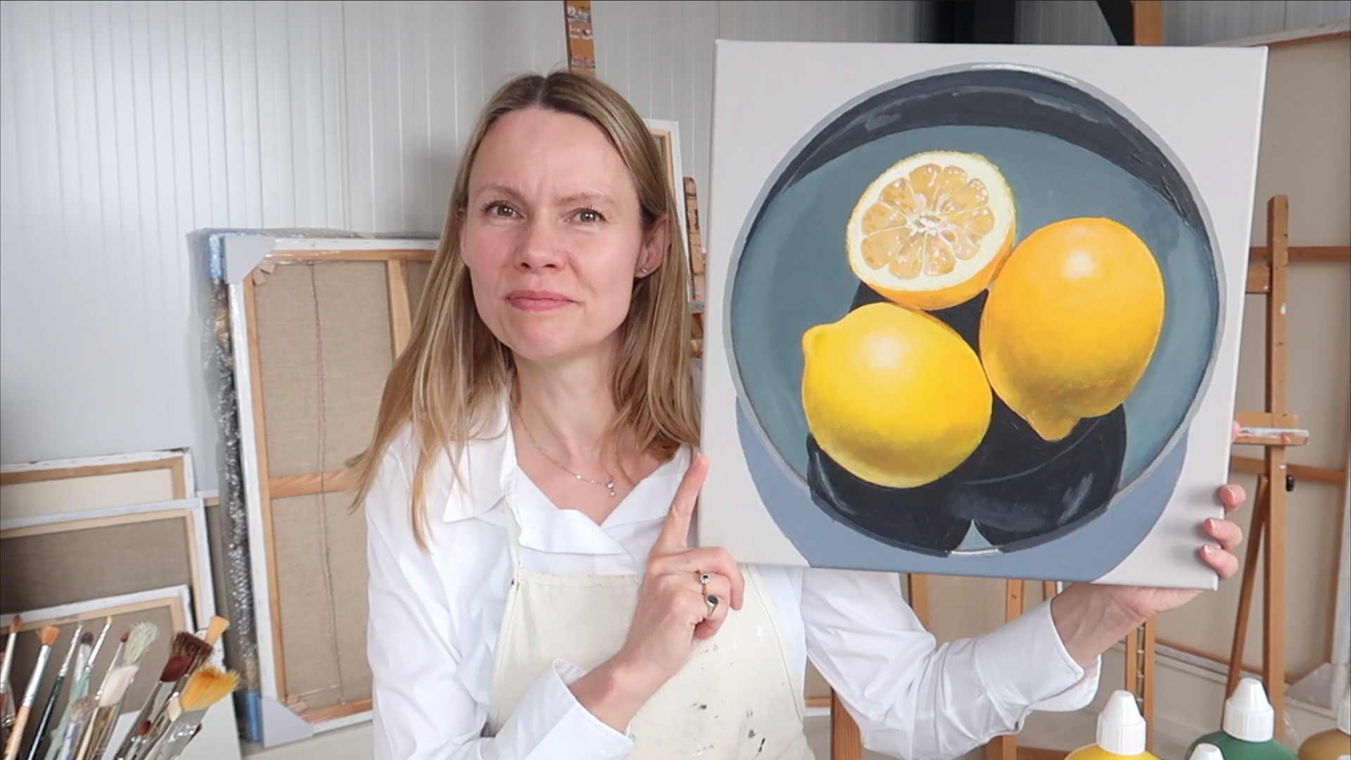Hannelore Houdijk in een witte blouse met een schilderij van citroenen en een doorgesneden citroen in haar handen in een atelier. Voor een online schildercursus.