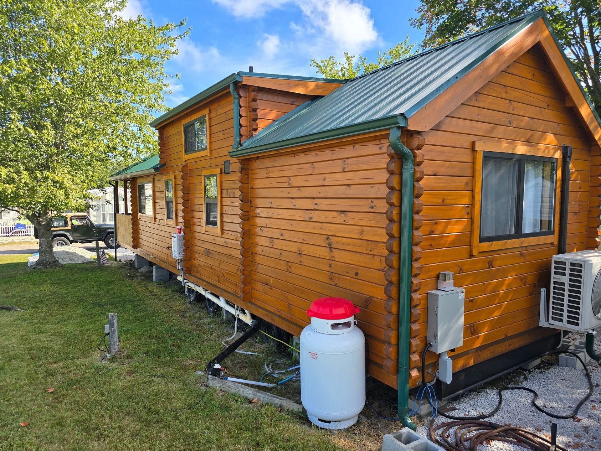 Wooden cabin exterior with green roof, trees nearby, and a white water barrel in front