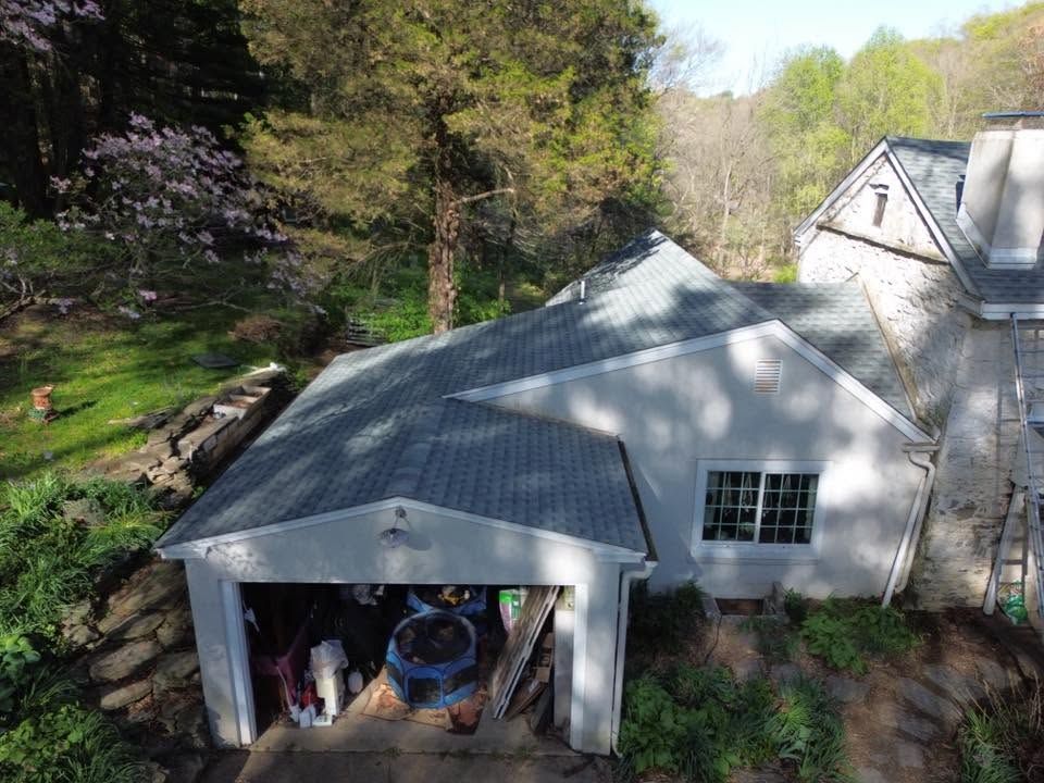 Aerial view of a gray-roofed garage beside a house, surrounded by trees and a sloped wooded yard.