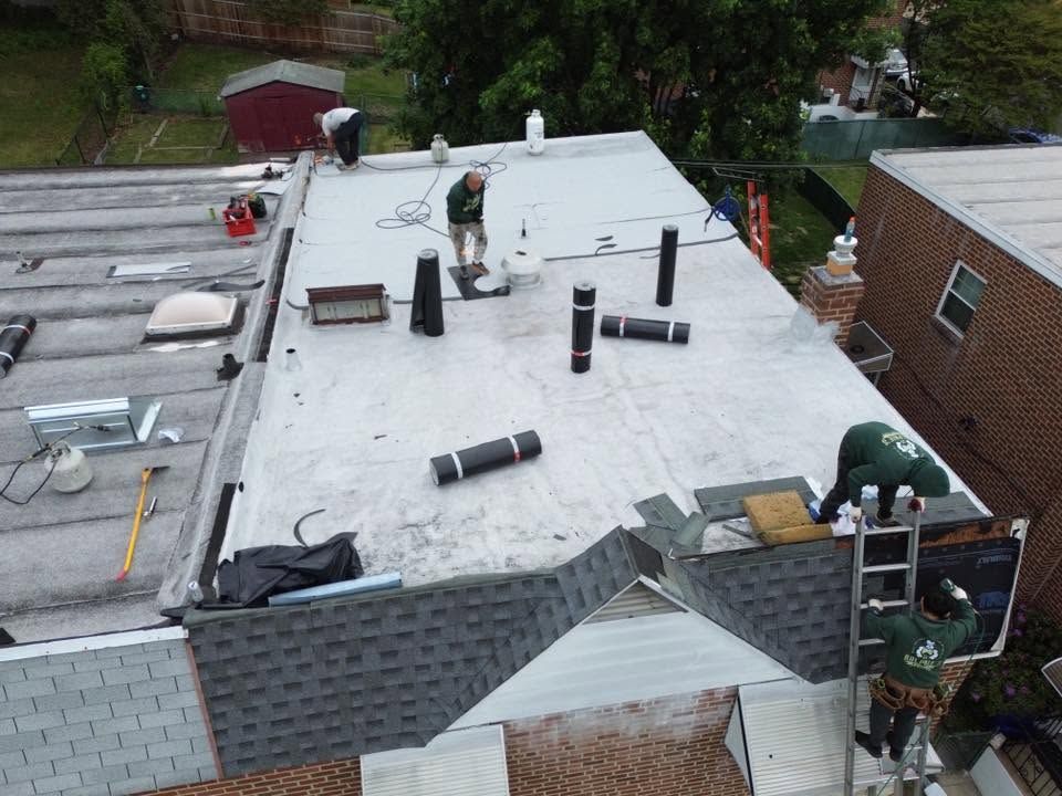 Roofers working on a flat white rooftop with vents and materials, beside a brick building.