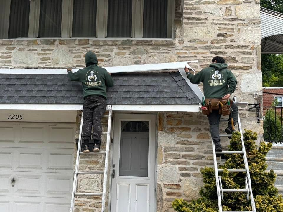 Two workers on ladders repair a stone house roof under a window, wearing green jackets.