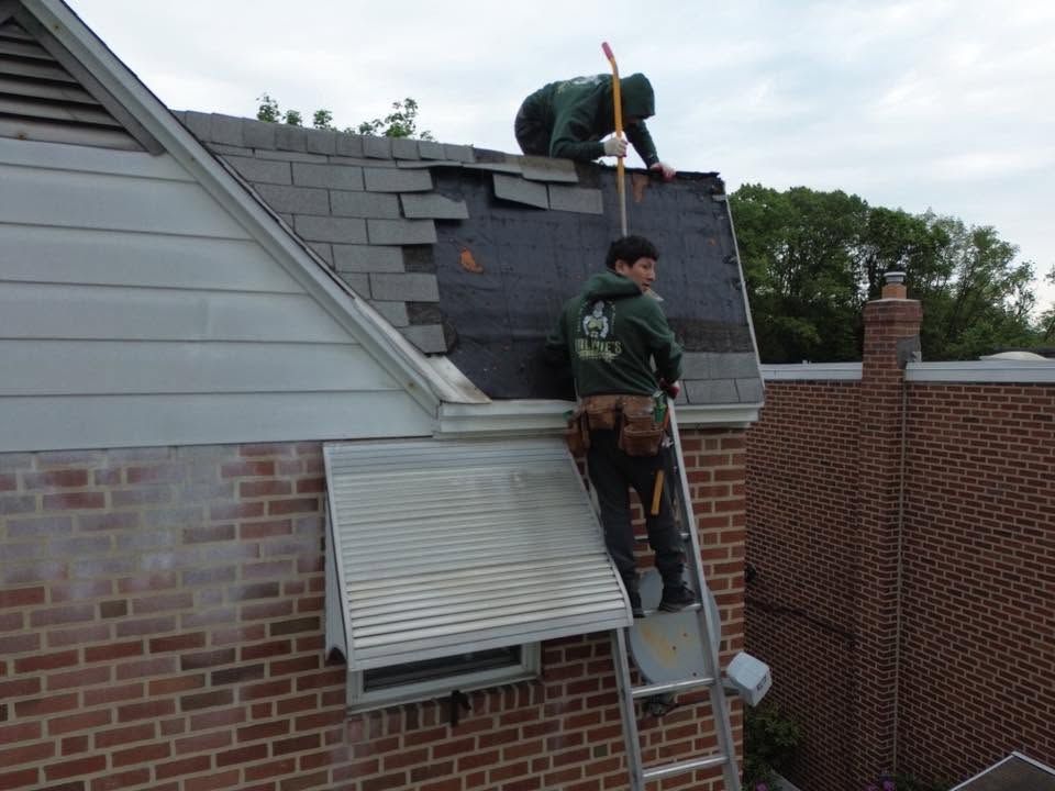 Workers repairing a roof, one on a ladder and one on the shingles beside a brick house