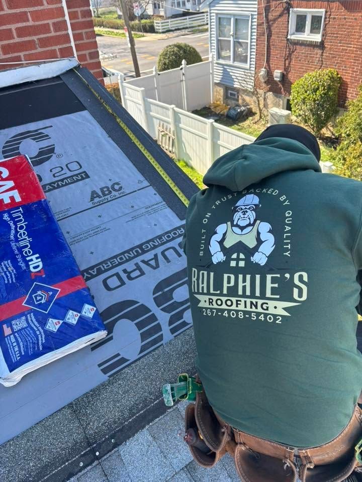 Roofing worker in green hoodie installing underlayment on a sloped roof beside a brick house.