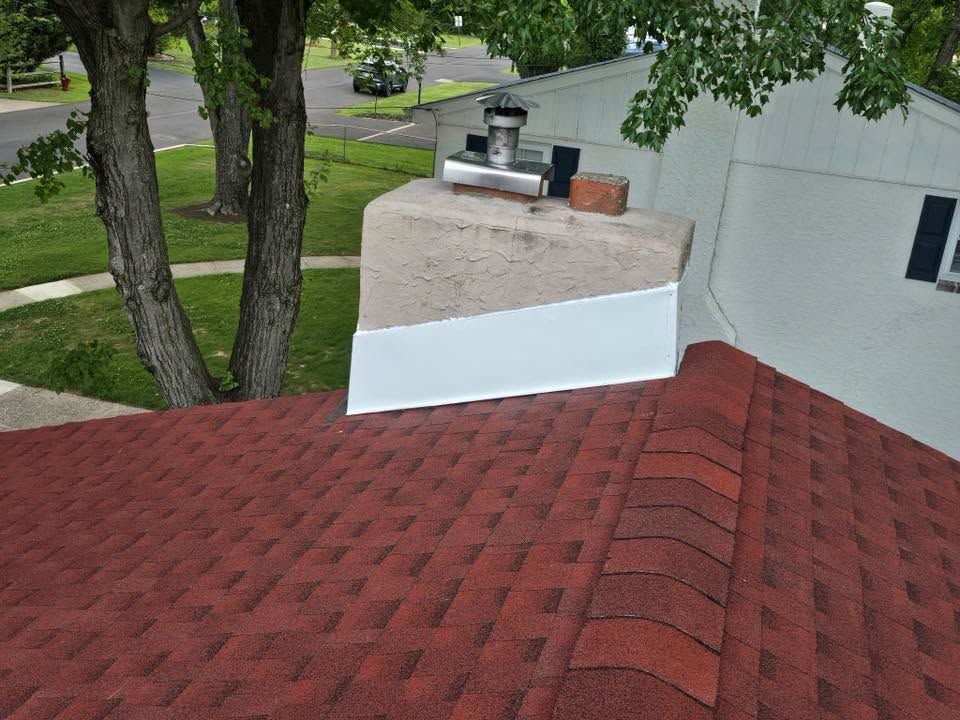 Red shingle roof with a white chimney and small vent near trees and a house wall