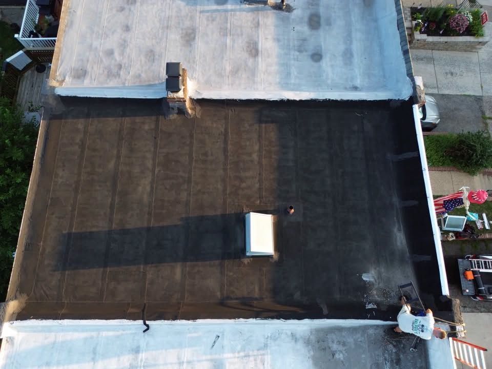 Aerial view of a flat rooftop with dark coating, skylight, and long shadow.