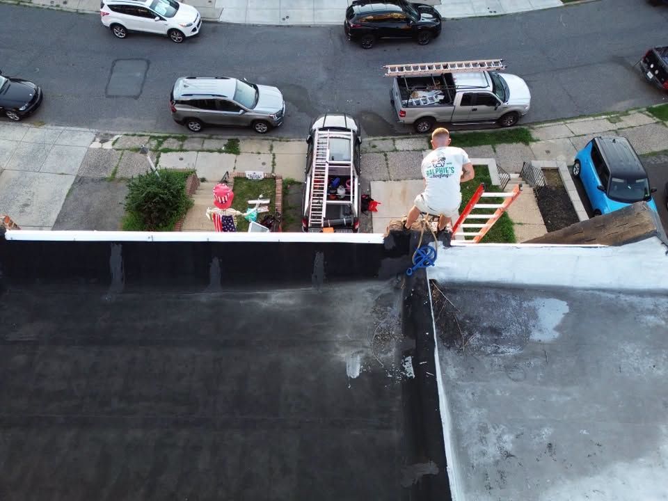 Person standing on a rooftop near the edge, looking down at a street with parked cars and a ladder below