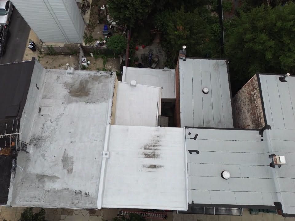 Aerial view of flat rooftops with white and gray surfaces beside trees and a narrow alley