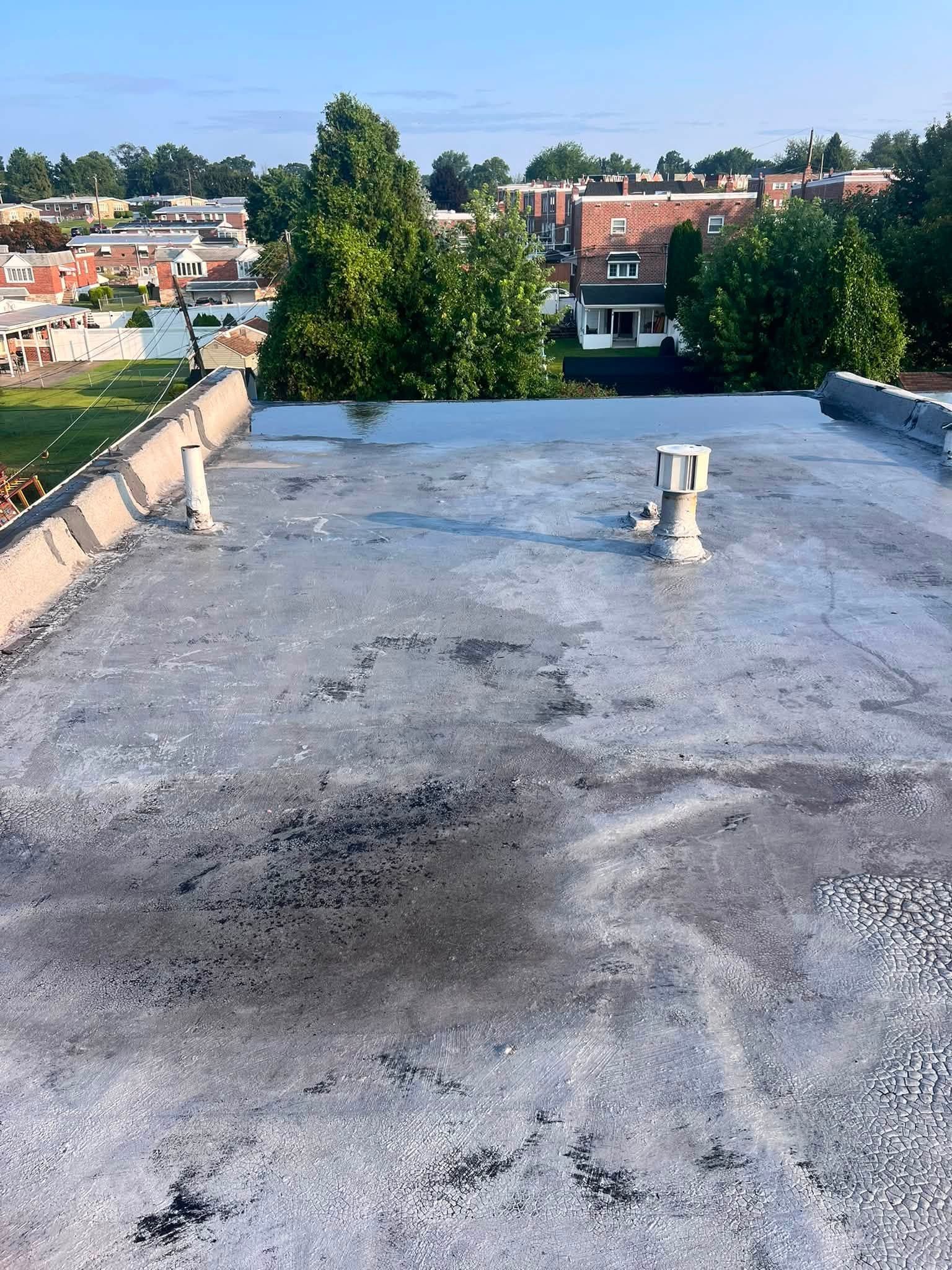 Rooftop with weathered gray surface, vents, and a city neighborhood visible beyond trees.
