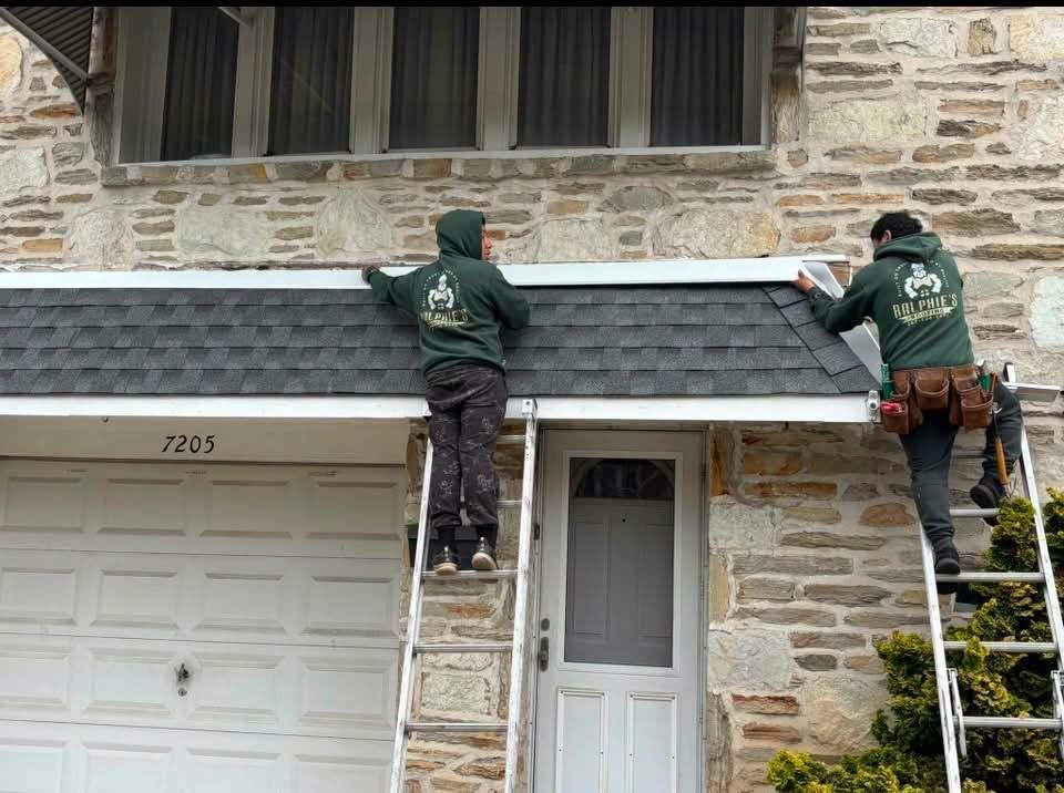 Two workers on ladders repairing a gray shingled roof above a stone house entrance