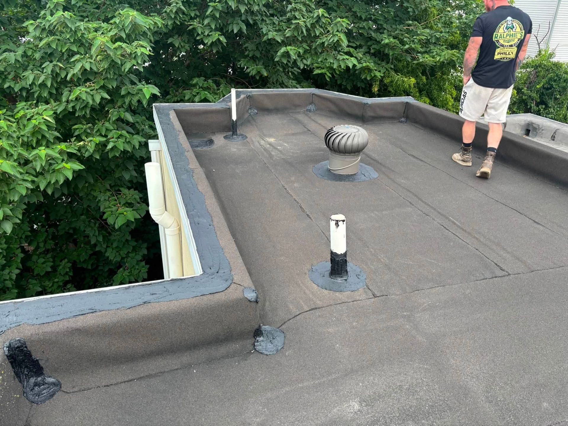 Flat rooftop with gravel, vents, and a person standing near the edge beside trees.