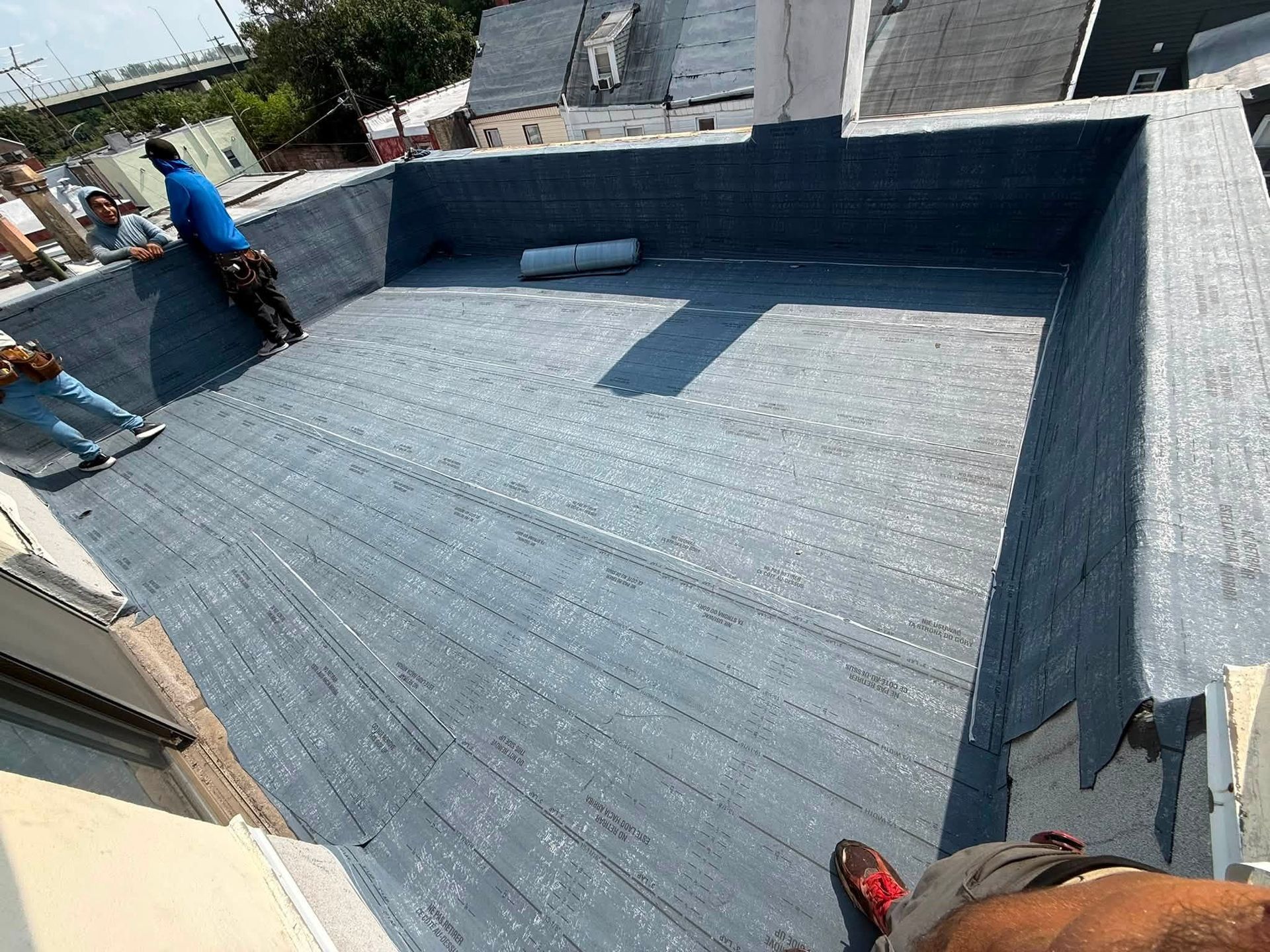 Workers installing blue waterproofing on a rooftop terrace with a low parapet wall