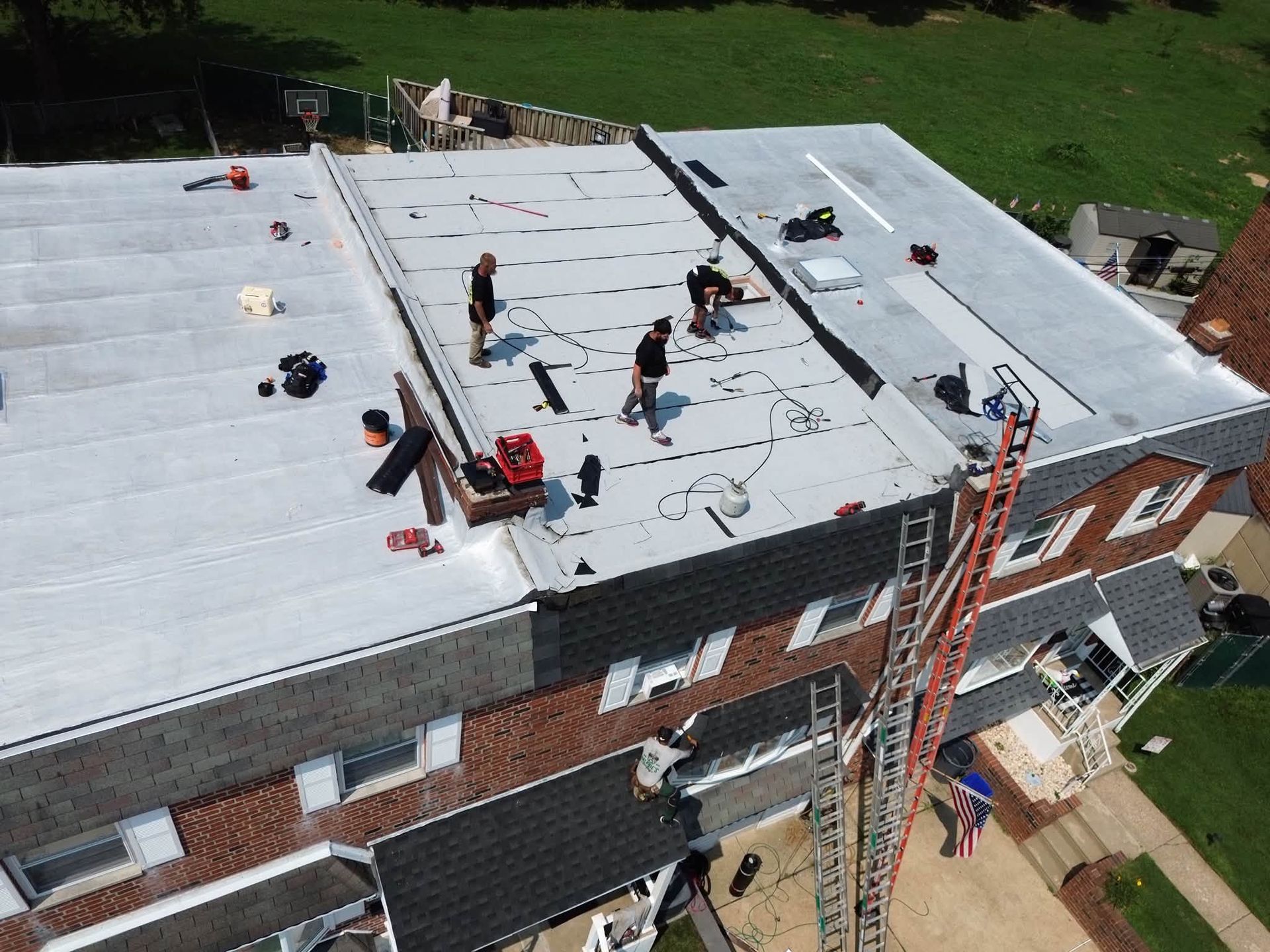 Workers on a flat rooftop installing a white roof on a brick building, with ladders and tools visible.