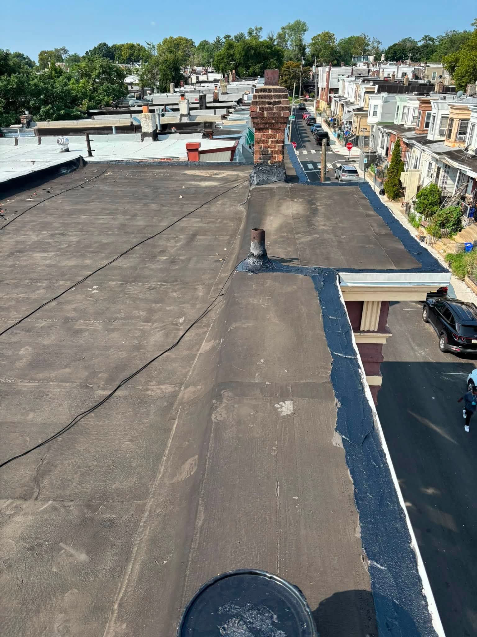 Rooftop with flat tar surface, chimney vent, and row of houses in the distance under a clear sky