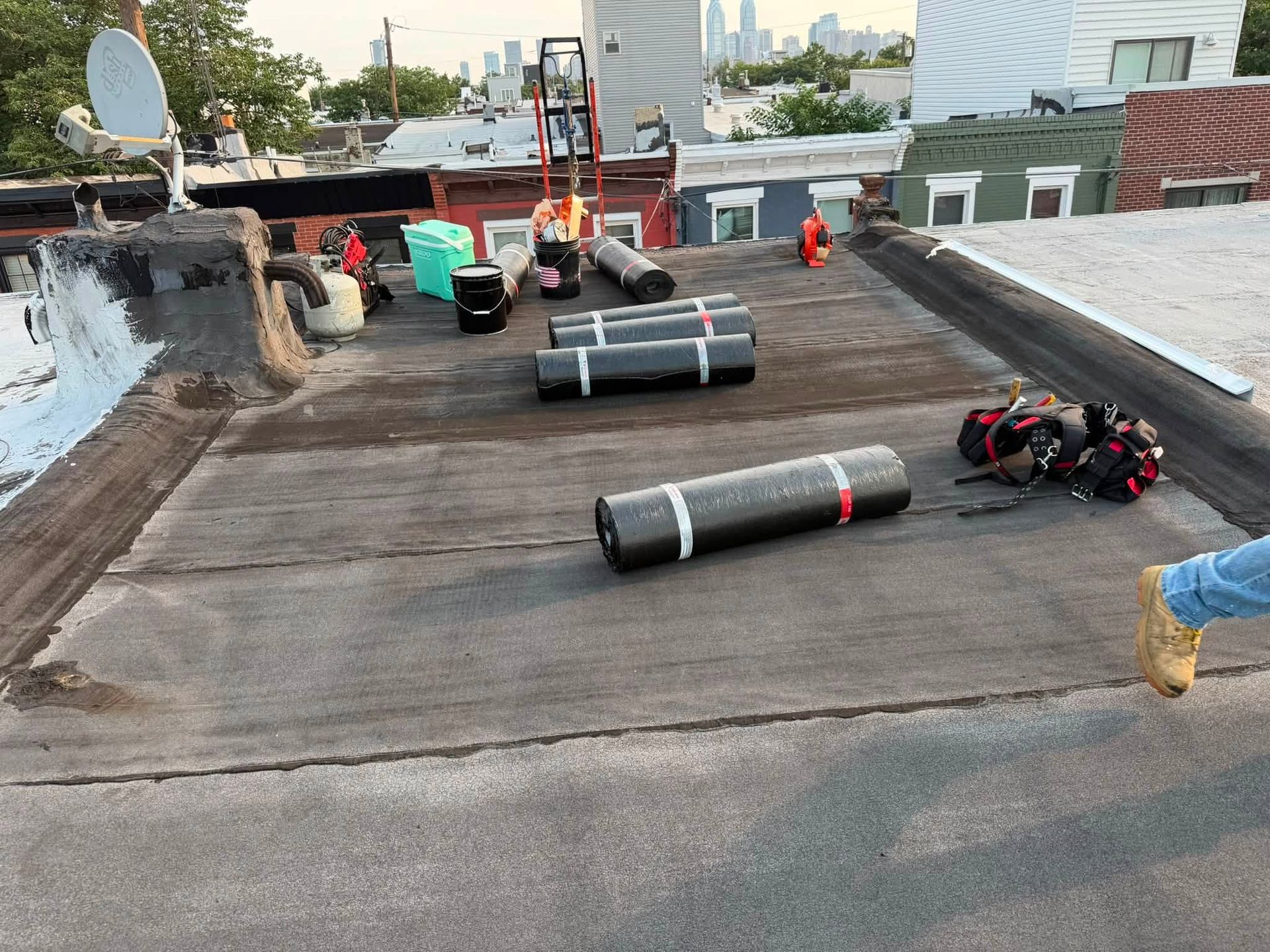 Rooftop with rolled black roofing material, tools, and scattered supplies near a parapet wall.