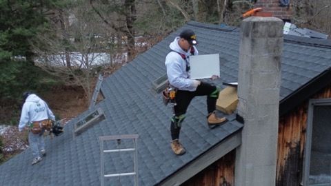Workers installing solar panels on a gray shingled roof with a chimney nearby.