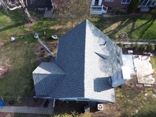 Aerial view of a house with a gray roof and small front porch, surrounded by yard and trees.