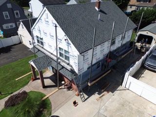 House under construction with white siding wrap, gray roof, and scaffolding on a driveway beside a lawn