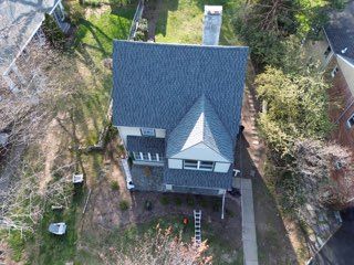 Aerial view of a gray house with a dark roof, chimney, and ladder in the front yard, surrounded by trees.