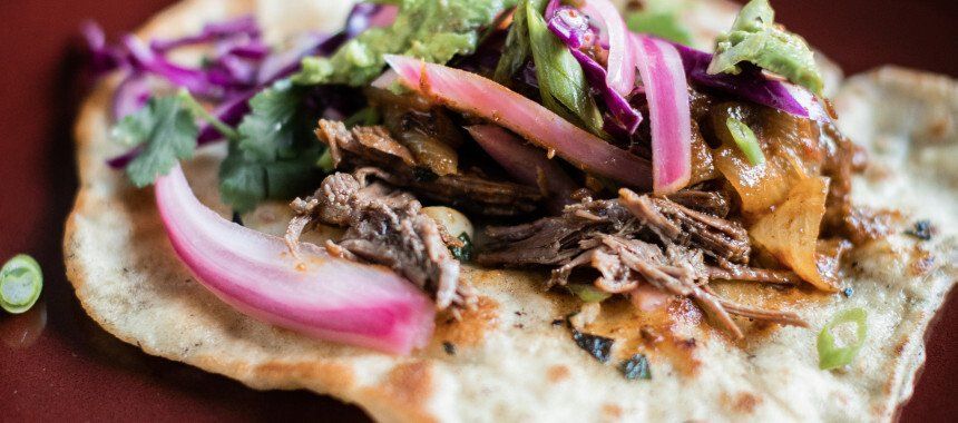 Taco topped with shredded meat, pickled onions, red cabbage, cilantro, and green onions.