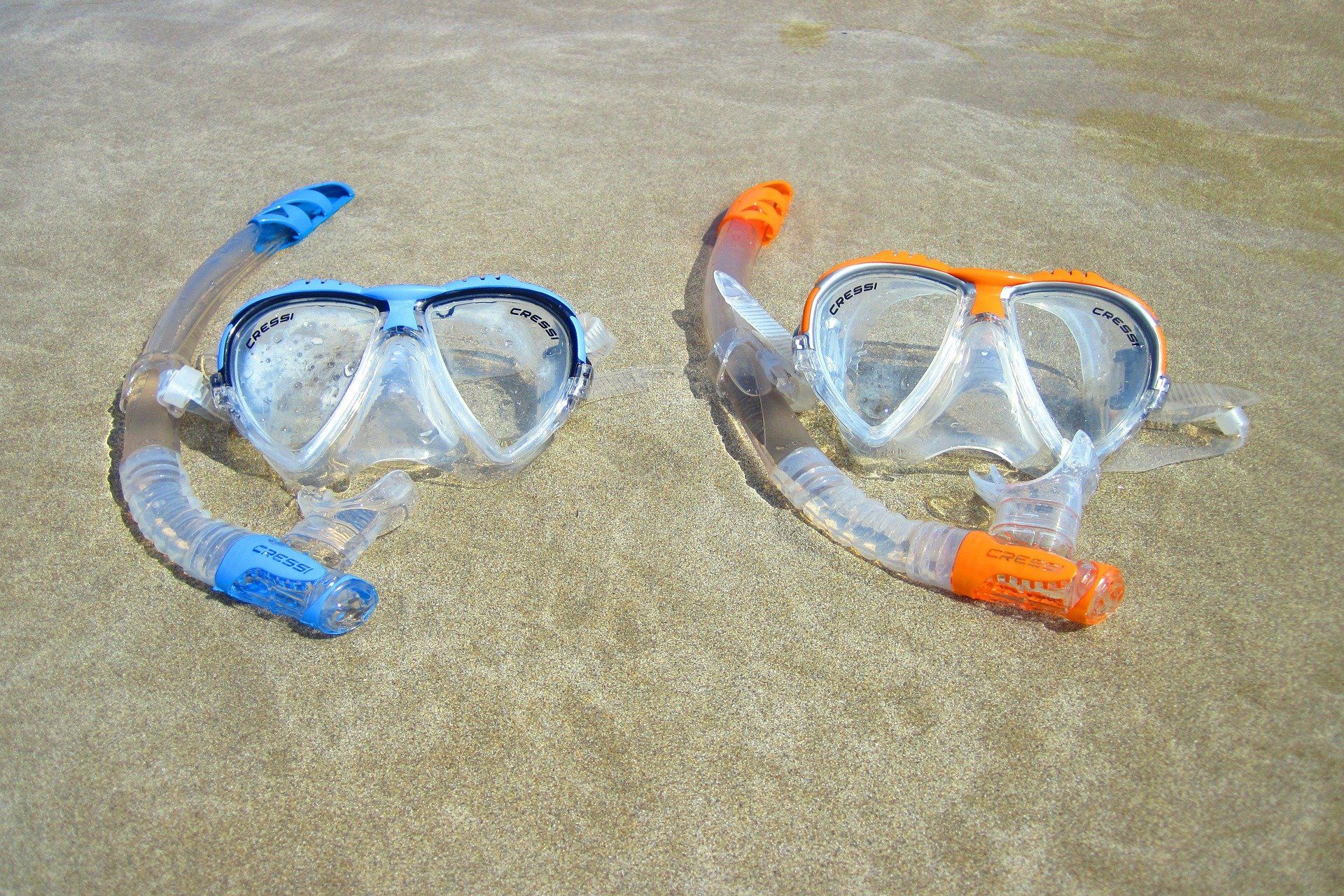 Two snorkel masks, blue and orange, on a sandy surface in shallow water.