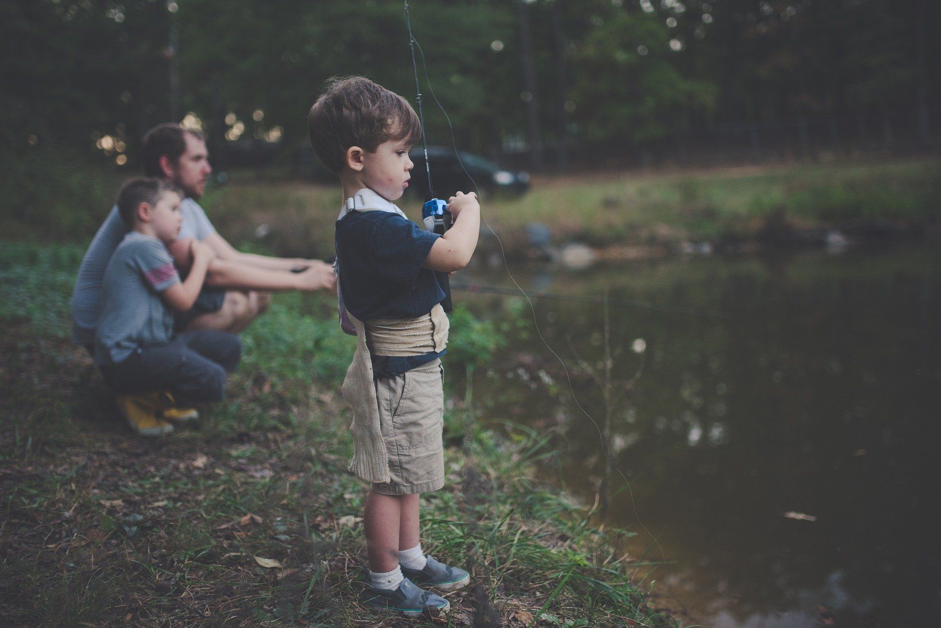 Boy fishing at a pond, with a man and another boy watching nearby. Dark green setting.