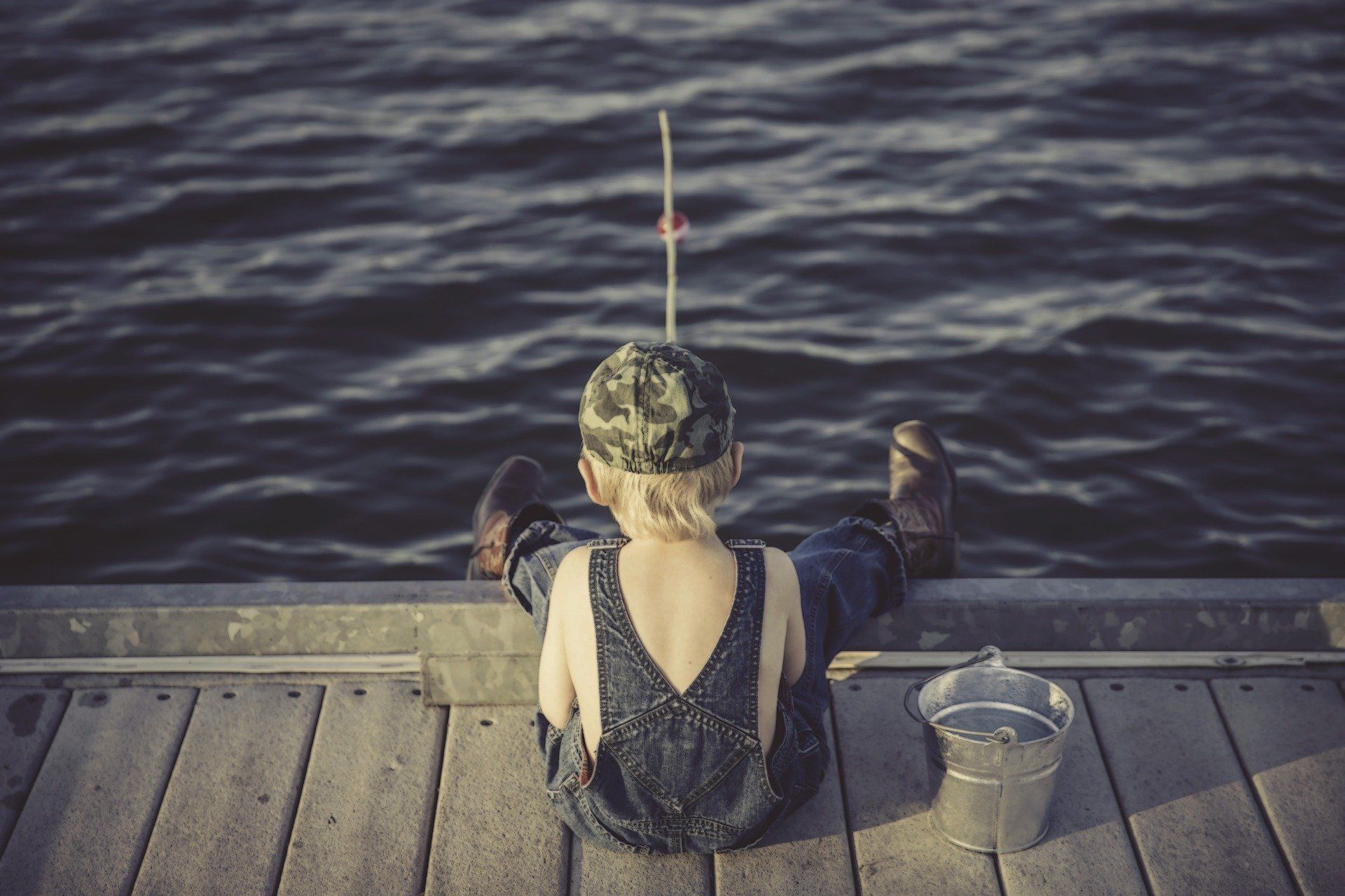 Child in overalls and camouflage hat fishing from a dock, facing the water. A bucket sits beside them.