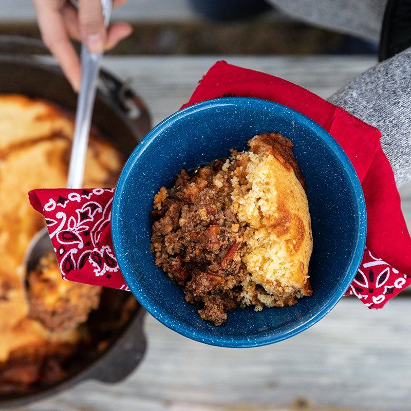 A person serving a portion of cornbread and ground meat casserole from a cast iron dish into a blue bowl.