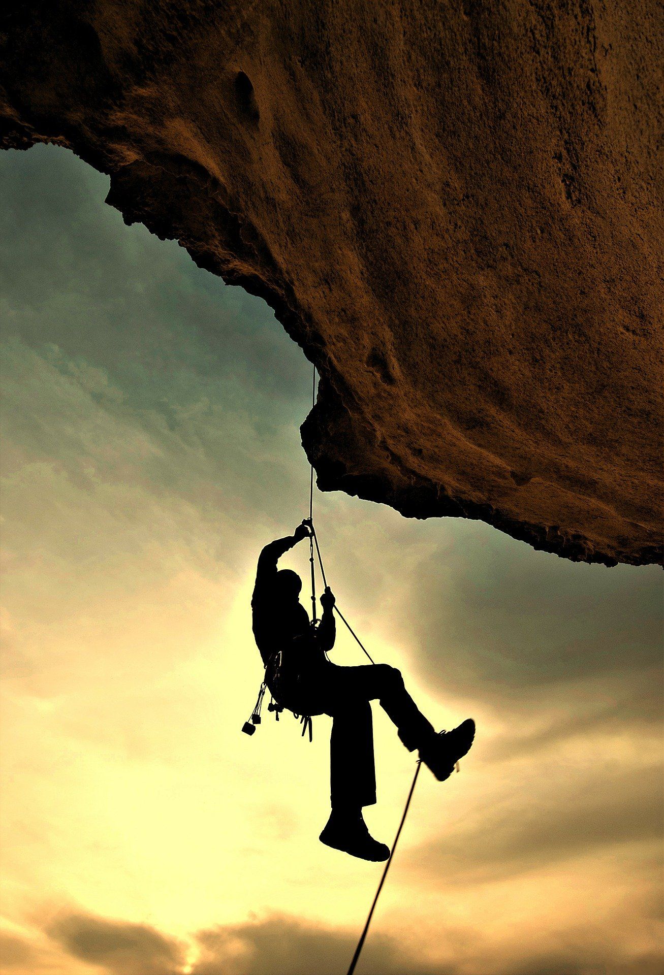 Silhouette of a person rappelling down a cliff face against a cloudy sky, rope visible.