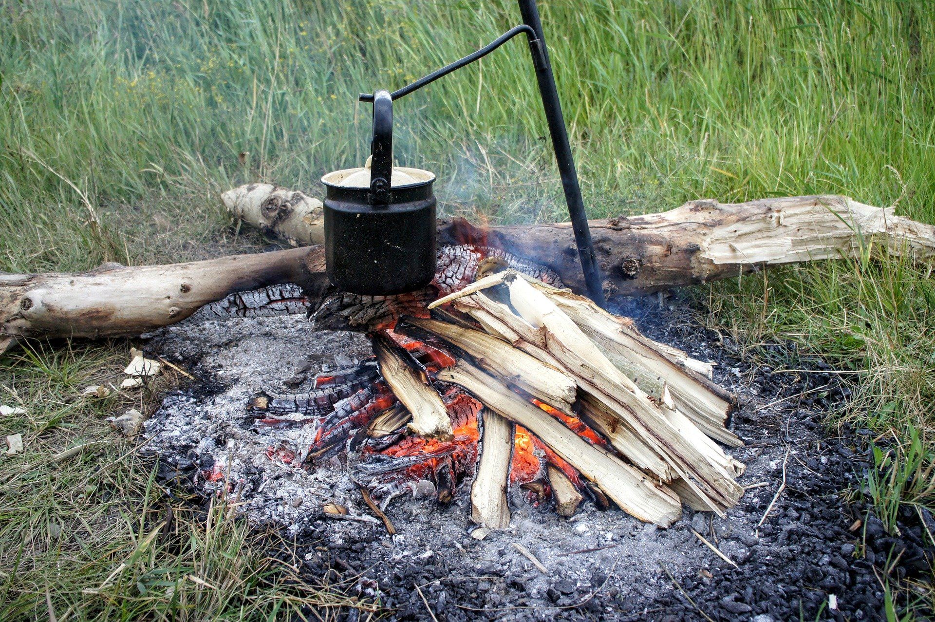 A campfire with a metal pot suspended above it, in a grassy setting.