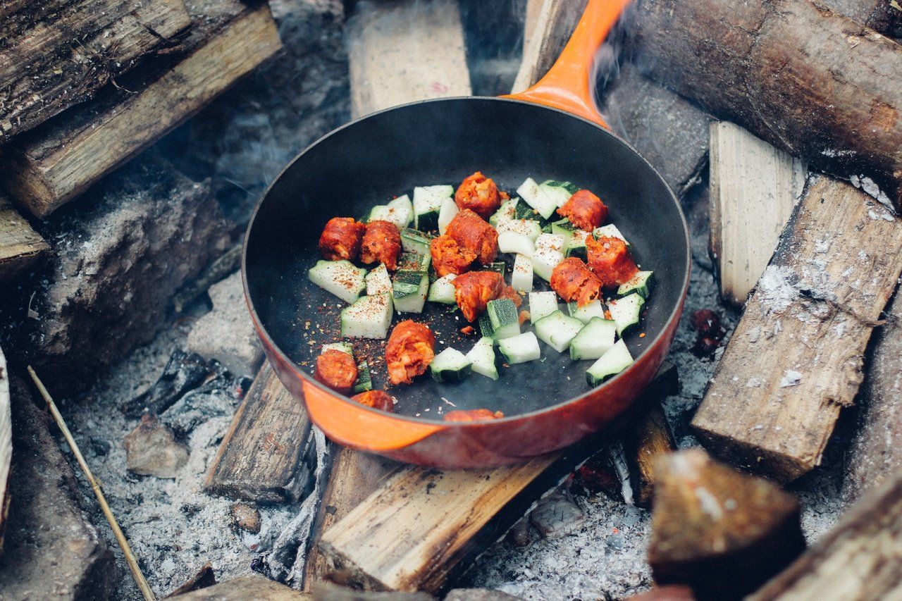 Frying pan with chorizo and zucchini cooking over campfire. Orange handle, wood, and smoke visible.