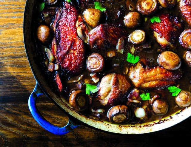 Chicken in a dark sauce with mushrooms, in a blue-handled pot on wood.