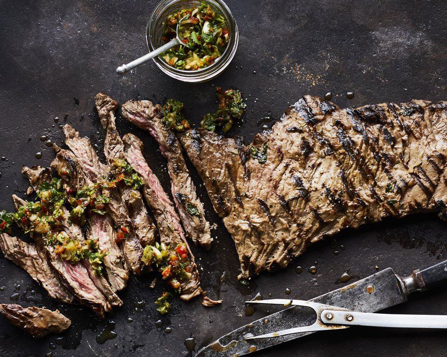 Grilled steak, sliced and topped with chimichurri sauce, on a dark surface, with a small bowl of sauce and a knife.