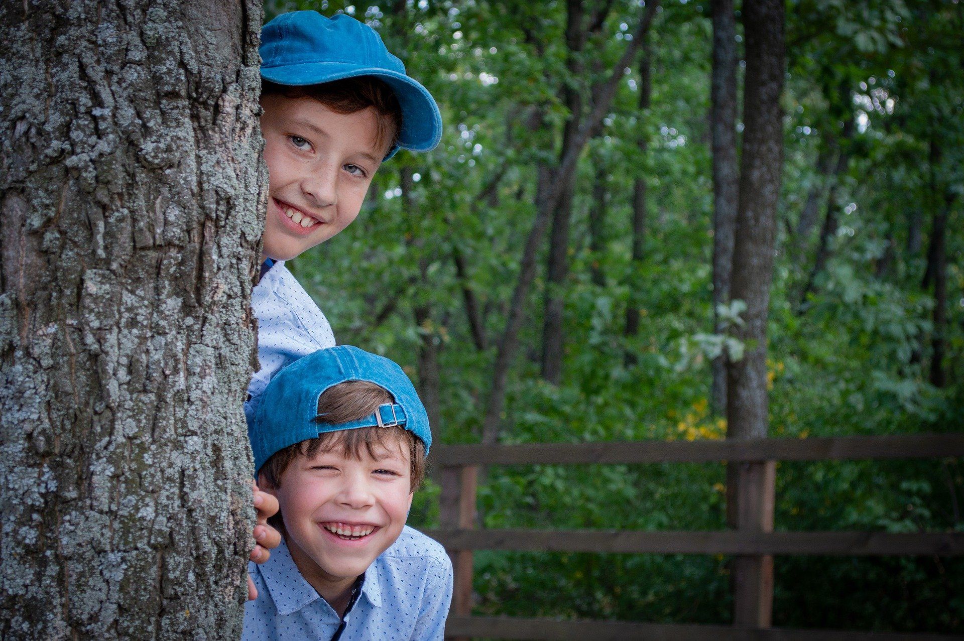 Two children peek around a tree trunk, smiling and wearing blue caps in a wooded area.
