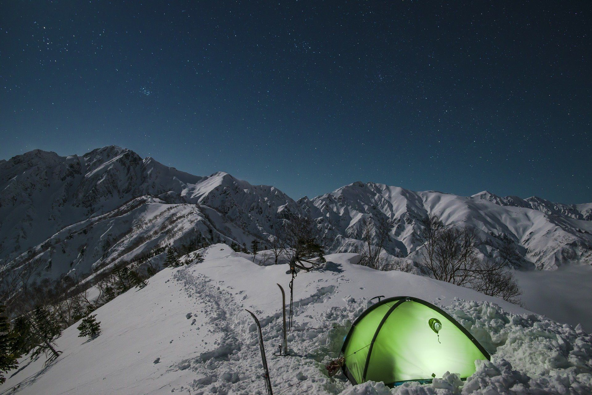 Snowy mountain range at night with a lit green tent, under a starry sky.